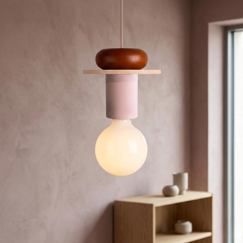 May include: A modern pendant light fixture with a large, round, illuminated bulb. The fixture features a wooden top, a light pink cylinder, and a wooden accent. The light hangs from a thin cord, showcasing a minimalist design.