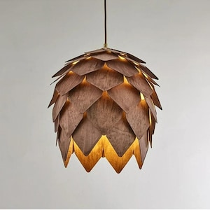 May include: A brown, pendant light fixture with a layered, geometric design. The fixture resembles a pinecone, with overlapping wooden panels. The interior glows with warm light, creating a cozy ambiance. The light hangs from a brown cord.