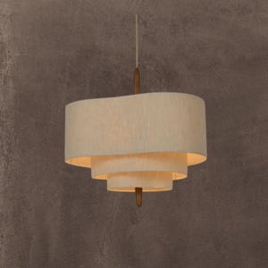 Peut inclure: Un luminaire suspendu superposé de couleur crème avec un accent en bois. La lampe comporte trois abat-jours ovales empilés, chacun légèrement plus petit que le précédent. Le luminaire est suspendu à un cordon fin et à une tige en bois.