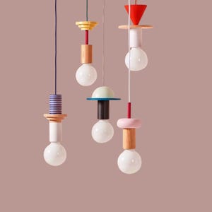 Peut inclure: Ensemble de lampes suspendues modernes avec des accents g&eacute;om&eacute;triques en bois color&eacute;. Chaque lampe comprend une ampoule blanche, suspendue par un cordon, et orn&eacute;e de formes rouges, roses, jaunes, bleues et en bois naturel. Le fond est rose p&acirc;le.