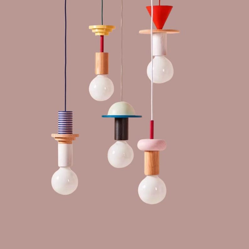 Peut inclure: Ensemble de lampes suspendues modernes avec des accents g&eacute;om&eacute;triques en bois color&eacute;. Chaque lampe comprend une ampoule blanche, suspendue par un cordon, et orn&eacute;e de formes rouges, roses, jaunes, bleues et en bois naturel. Le fond est rose p&acirc;le.
