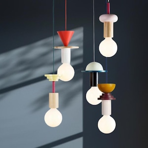 Puede incluir: Una colección de lámparas colgantes modernas con formas geométricas coloridas. Cada lámpara presenta una bombilla blanca, elementos de madera y una variedad de acentos de color, incluyendo rojo, amarillo y azul. Las luces cuelgan de cables de colores.