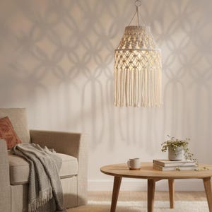 Puede incluir: Lámpara colgante de macramé color crema, de forma cilíndrica con flecos. La luz cuelga de un anillo de metal y proyecta una sombra decorativa en la pared. Está suspendida sobre una mesa de centro redonda de madera.