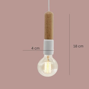 Peut inclure: Un luminaire suspendu avec une partie sup&eacute;rieure en bois, une section centrale en c&eacute;ramique blanche et une ampoule transparente. L'ampoule mesure 4 cm de large et le luminaire 18 cm de long. Il est suspendu &agrave; un cordon blanc sur fond rose.