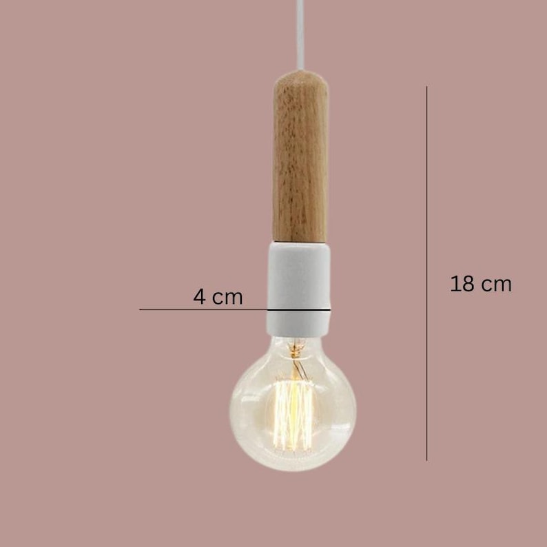 Peut inclure: Un luminaire suspendu avec une partie sup&eacute;rieure en bois, une section centrale en c&eacute;ramique blanche et une ampoule transparente. L'ampoule mesure 4 cm de large et le luminaire 18 cm de long. Il est suspendu &agrave; un cordon blanc sur fond rose.