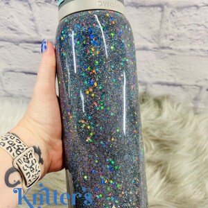 May include: A black Owala water bottle covered in iridescent glitter. The bottle has a gray lid with a black spout and a teal button. The bottle is held in a hand with a leopard print watch band. The text "Kritters Kreations" is visible.