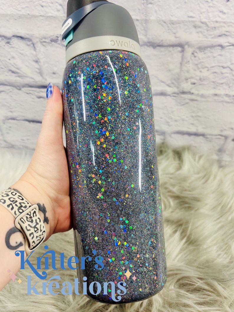 May include: A black Owala water bottle covered in iridescent glitter. The bottle has a gray lid with a black spout and a teal button. The bottle is held in a hand with a leopard print watch band. The text "Kritters Kreations" is visible.