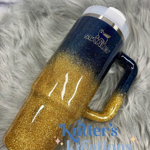 Puede incluir: Un termo de viaje Stanley con asa, con un dise&ntilde;o degradado de purpurina dorada en la parte inferior que se desvanece en purpurina azul oscuro en la parte superior. El termo tiene una tapa blanca y el logotipo de Stanley en dorado.