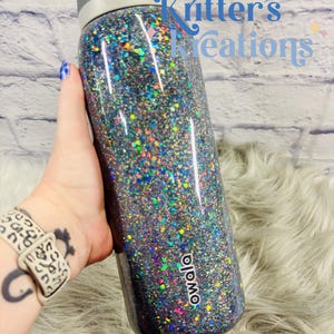May include: A glitter-covered Owala water bottle with a gray lid and a black base. The bottle is covered in iridescent glitter that reflects multiple colors. The text "Kritter's Creations" is visible in the background.