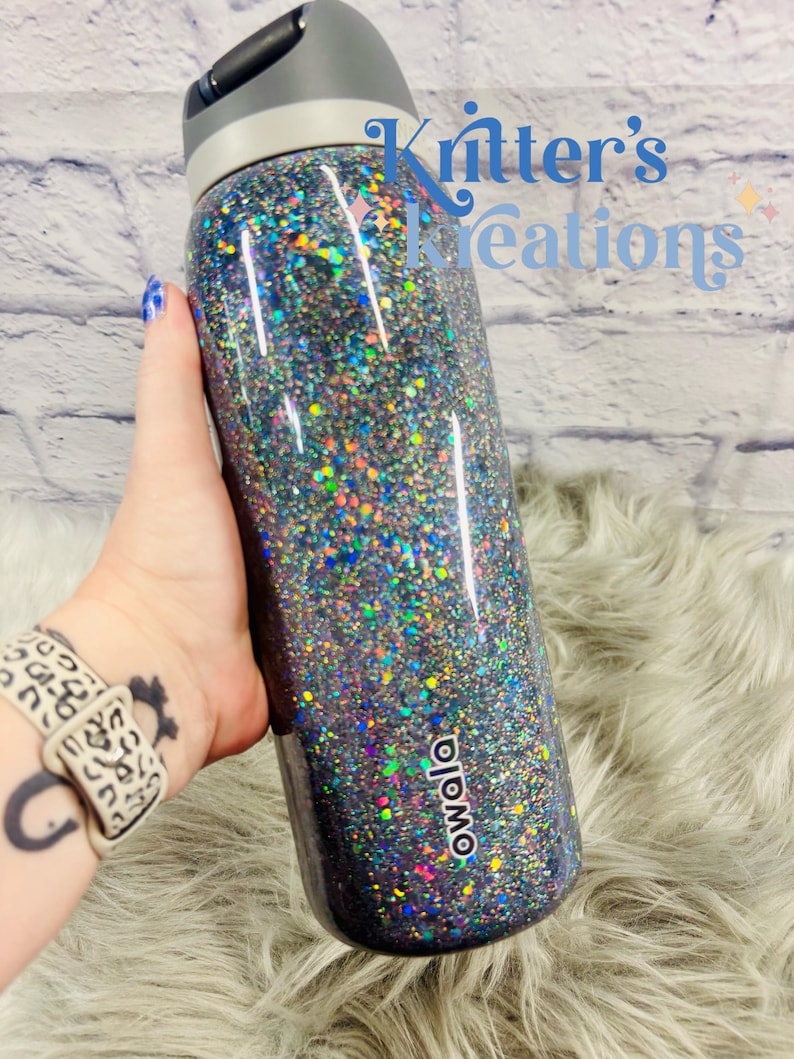 May include: A glitter-covered Owala water bottle with a gray lid and a black base. The bottle is covered in iridescent glitter that reflects multiple colors. The text "Kritter's Creations" is visible in the background.