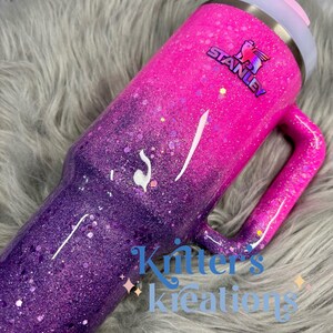 May include: A glittery Stanley tumbler in a gradient of pink and purple. The tumbler has a handle and a light gray lid with a pink slider. The Stanley logo is visible. The text "Kritter's Kreations" is at the bottom.