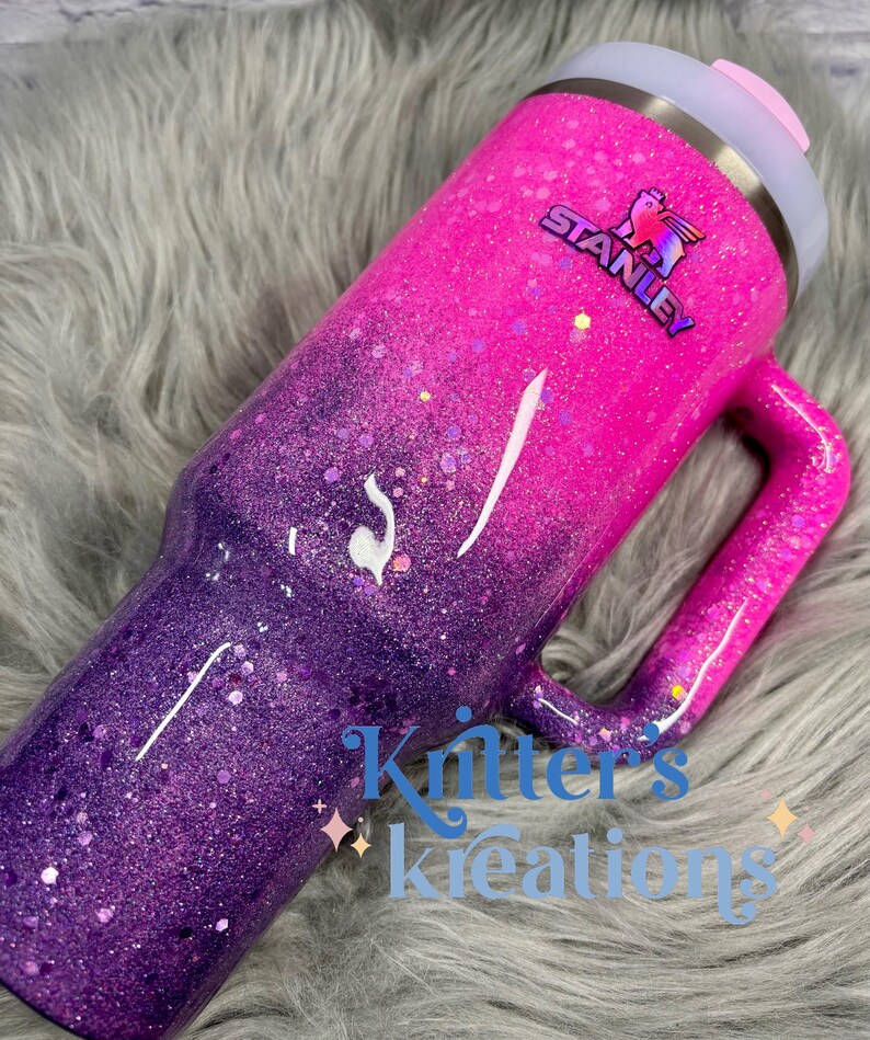 May include: A glittery Stanley tumbler in a gradient of pink and purple. The tumbler has a handle and a light gray lid with a pink slider. The Stanley logo is visible. The text "Kritter's Kreations" is at the bottom.