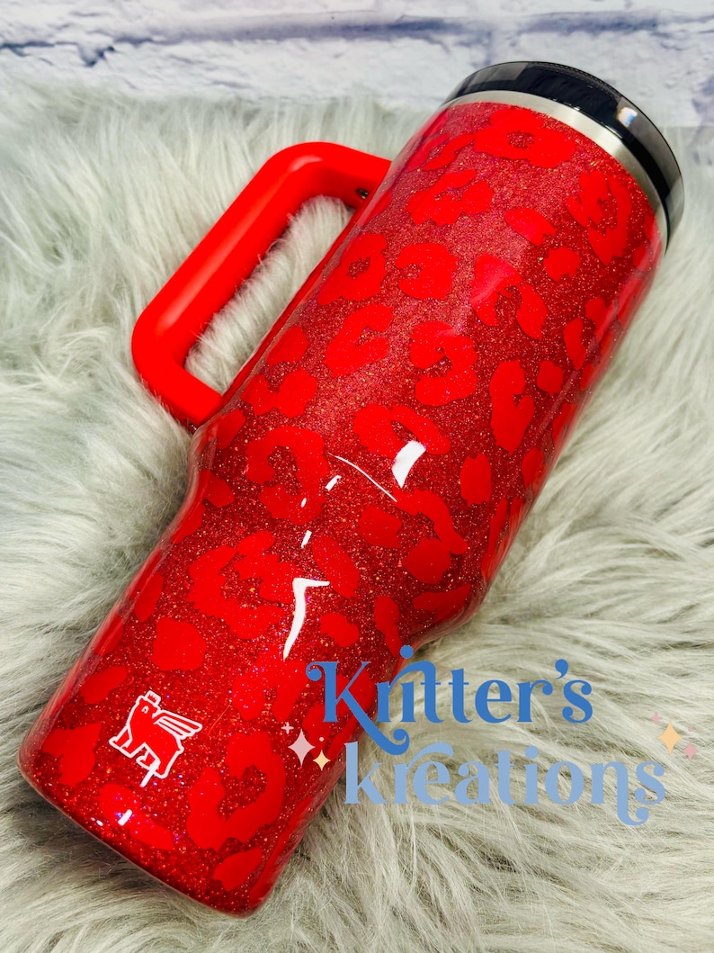 May include: A red glitter tumbler with a leopard print design and a red handle. The tumbler has a black lid and a white logo. The text "Kitter's Kreations" is visible on the side.