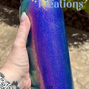 Peut inclure: Une bouteille d'eau cylindrique Owala avec un couvercle noir et une finition scintillante multicolore vibrante qui passe par des nuances de bleu, de violet et de vert. Le texte "Kritter's Kreations" est imprim&eacute; en bleu sur la partie sup&eacute;rieure.