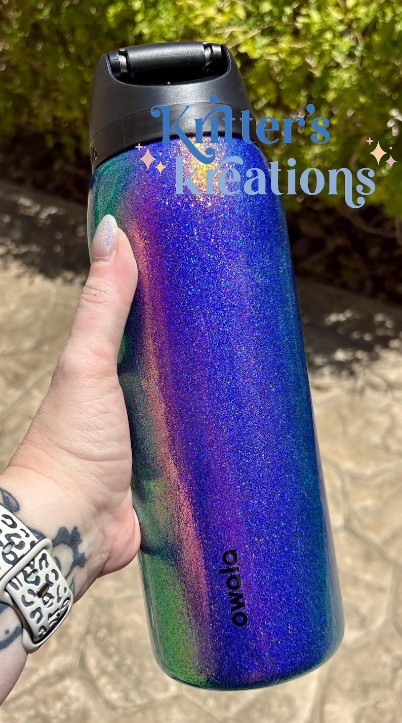 Peut inclure: Une bouteille d'eau cylindrique Owala avec un couvercle noir et une finition scintillante multicolore vibrante qui passe par des nuances de bleu, de violet et de vert. Le texte "Kritter's Kreations" est imprim&eacute; en bleu sur la partie sup&eacute;rieure.