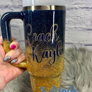 Puede incluir: Vaso con purpurina con un dise&ntilde;o degradado de azul marino a dorado. El vaso tiene un asa y una tapa blanca con pajita. Las palabras "Coach Kayla" est&aacute;n escritas en letras doradas. La parte inferior del vaso tiene el texto "Kitter's Kreations".