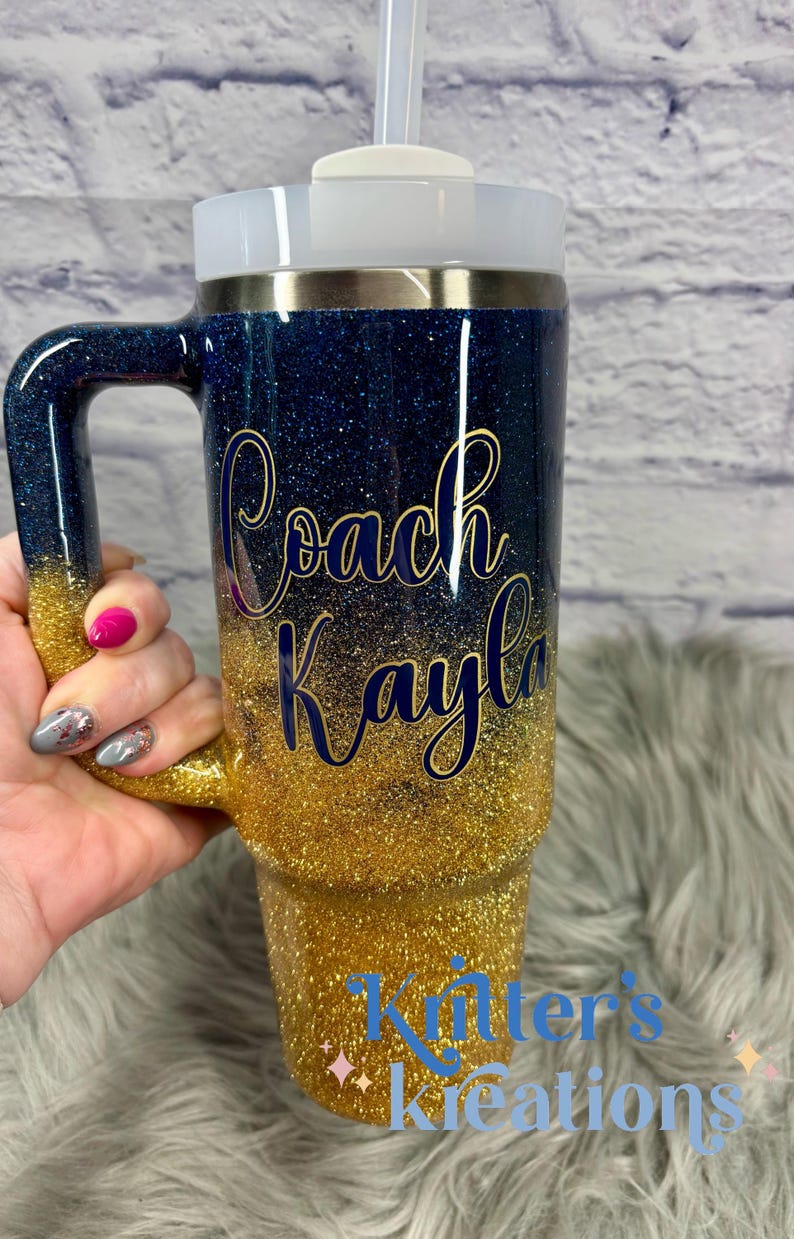Puede incluir: Vaso con purpurina con un dise&ntilde;o degradado de azul marino a dorado. El vaso tiene un asa y una tapa blanca con pajita. Las palabras "Coach Kayla" est&aacute;n escritas en letras doradas. La parte inferior del vaso tiene el texto "Kitter's Kreations".