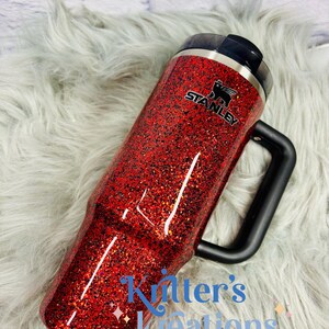 May include: A red glitter Stanley tumbler with a black handle and lid. The tumbler has the Stanley logo and the text "Kitter's Kreations" in blue script. The tumbler is on a fluffy, light gray surface.