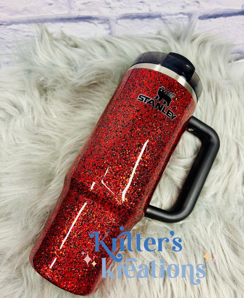 May include: A red glitter Stanley tumbler with a black handle and lid. The tumbler has the Stanley logo and the text "Kitter's Kreations" in blue script. The tumbler is on a fluffy, light gray surface.