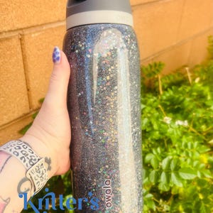 May include: A black glitter water bottle with a gray lid and handle. The bottle has the brand name "owala" printed on it. The text "Kritter's Kreations" is visible on the bottle.