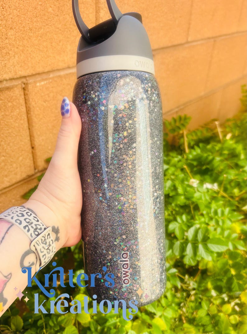 May include: A black glitter water bottle with a gray lid and handle. The bottle has the brand name "owala" printed on it. The text "Kritter's Kreations" is visible on the bottle.