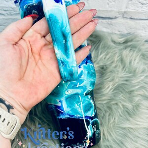 May include: A blue, white, and black tumbler with a swirl design. The tumbler has a handle and is made of stainless steel. The tumbler is on a fuzzy, light blue surface.