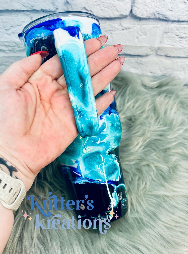 May include: A blue, white, and black tumbler with a swirl design. The tumbler has a handle and is made of stainless steel. The tumbler is on a fuzzy, light blue surface.