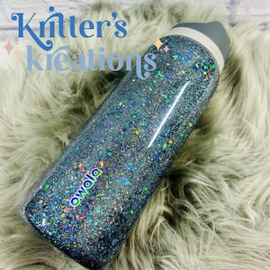 May include: A blue glitter Owala water bottle with a gray lid. The bottle is covered in iridescent glitter and has the text "Owala" in a colorful font. The text "Kritter's Kreations" is in a blue script font on the upper left.