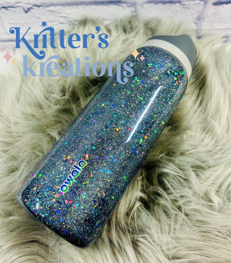 May include: A blue glitter Owala water bottle with a gray lid. The bottle is covered in iridescent glitter and has the text "Owala" in a colorful font. The text "Kritter's Kreations" is in a blue script font on the upper left.