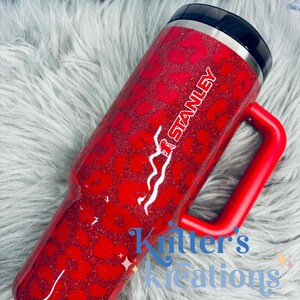 May include: Red glitter tumbler with a leopard print design. The tumbler has a red handle and the word "STANLEY" in white. The text "Kritter's Kreations" is also visible. The tumbler has a black lid.