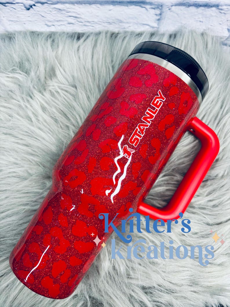 May include: Red glitter tumbler with a leopard print design. The tumbler has a red handle and the word "STANLEY" in white. The text "Kritter's Kreations" is also visible. The tumbler has a black lid.
