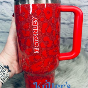May include: Red glitter Stanley tumbler with a leopard print design. Features a red handle and a black lid. The Stanley logo is visible on the side. The text "Kritter's Kreations" is also visible on the tumbler.