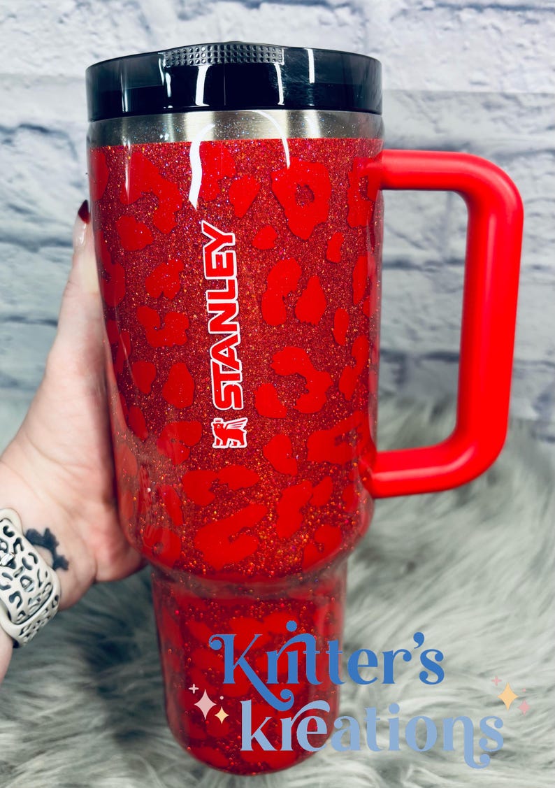 May include: Red glitter Stanley tumbler with a leopard print design. Features a red handle and a black lid. The Stanley logo is visible on the side. The text "Kritter's Kreations" is also visible on the tumbler.