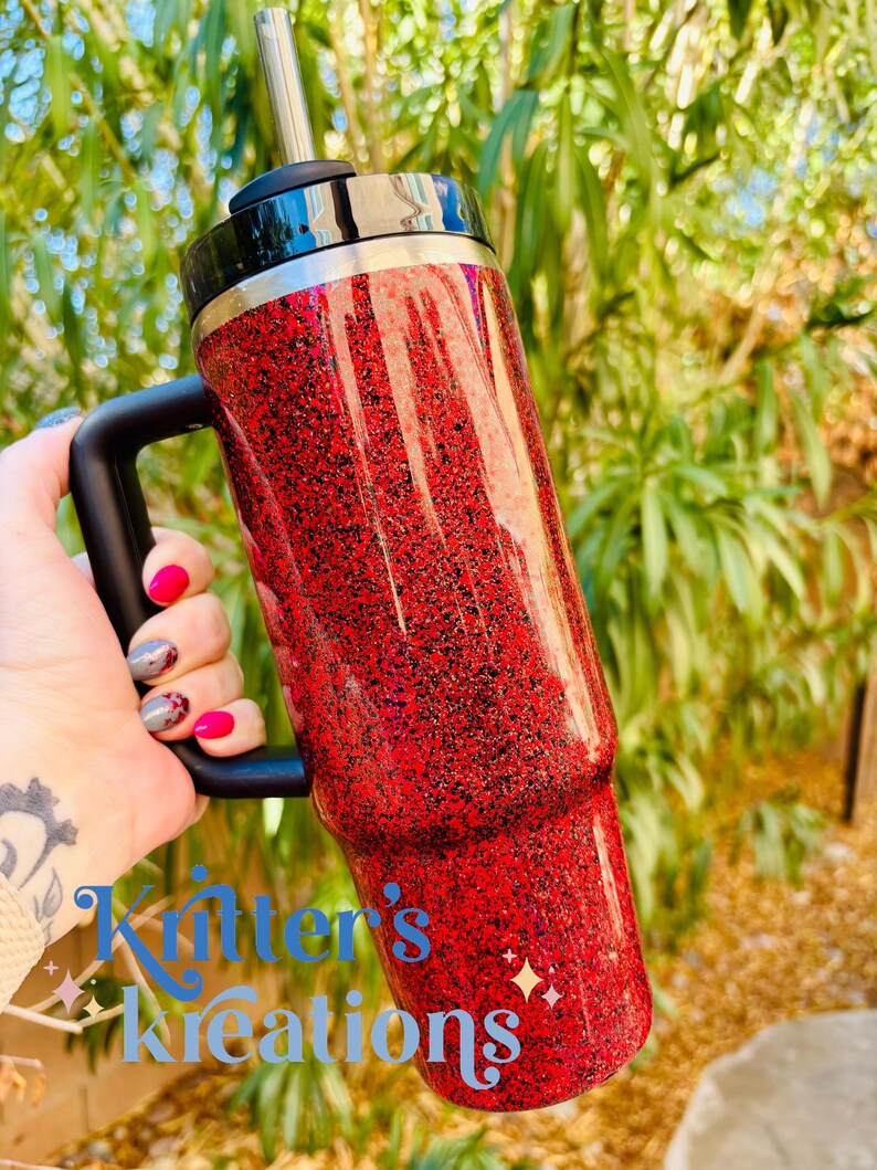 May include: A red glitter tumbler with a black handle and lid. The tumbler has a silver straw and a black rim. The text "Kitter's Kreations" is visible at the bottom of the tumbler.