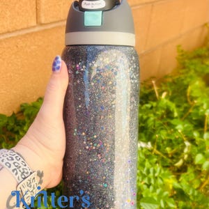 May include: A glitter-covered water bottle with a black and gray lid. The bottle is filled with a dark glitter and has a push button on the lid. The text "Push Button" is visible on the lid.