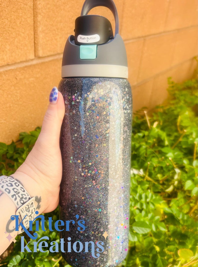 May include: A glitter-covered water bottle with a black and gray lid. The bottle is filled with a dark glitter and has a push button on the lid. The text "Push Button" is visible on the lid.