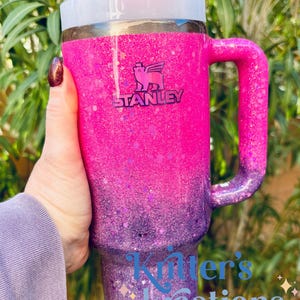 May include: A Stanley tumbler with a pink and purple glitter ombre design. The tumbler has a handle, a white lid, and a straw. The Stanley logo is visible. The text "Kitter's Kreations" is on the tumbler.