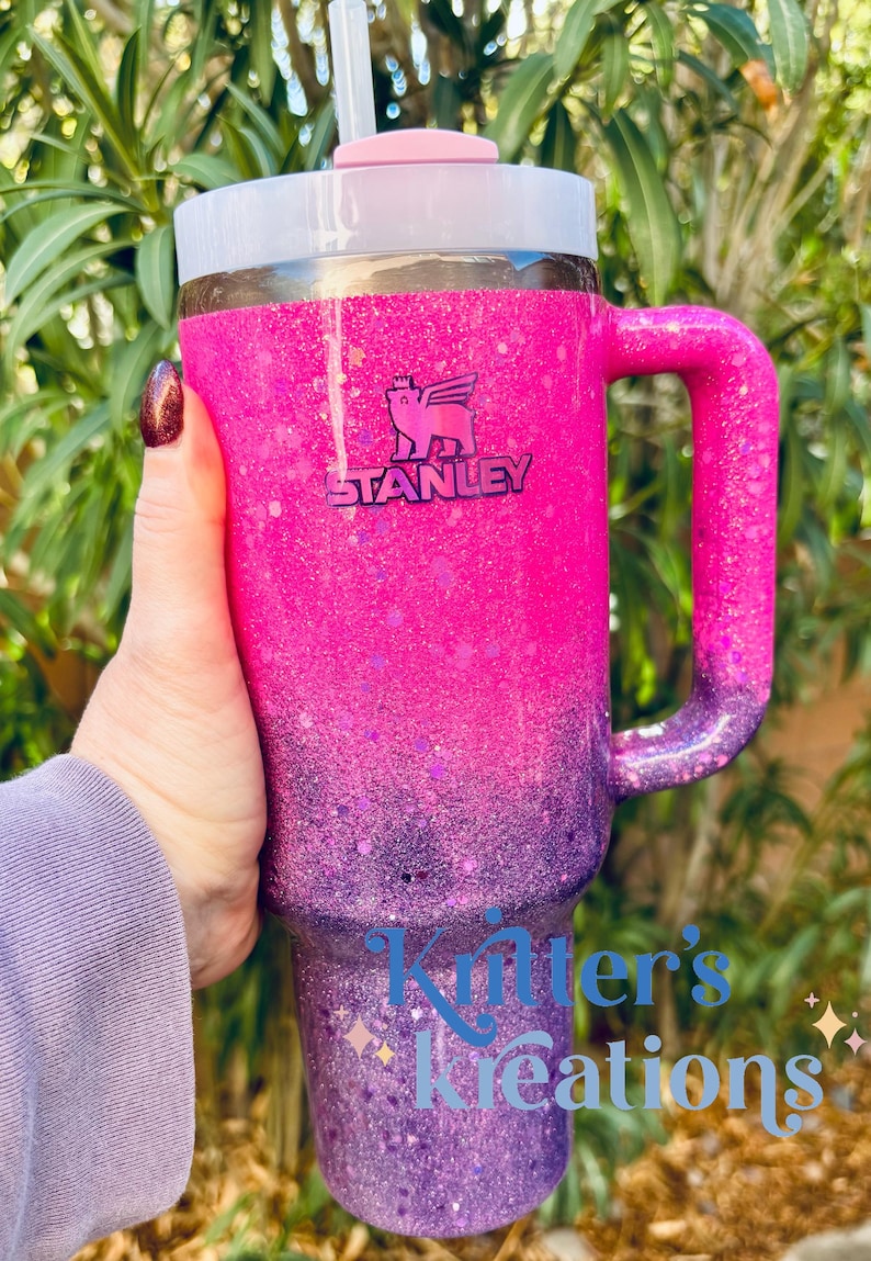 May include: A Stanley tumbler with a pink and purple glitter ombre design. The tumbler has a handle, a white lid, and a straw. The Stanley logo is visible. The text "Kitter's Kreations" is on the tumbler.