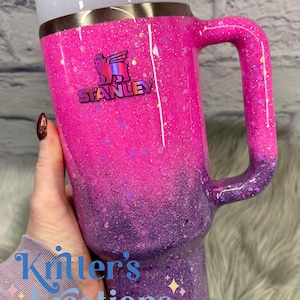 May include: A glittery Stanley tumbler in shades of pink and purple, with a handle and a straw. The tumbler features the Stanley logo and the text "Kitter's Kreations".