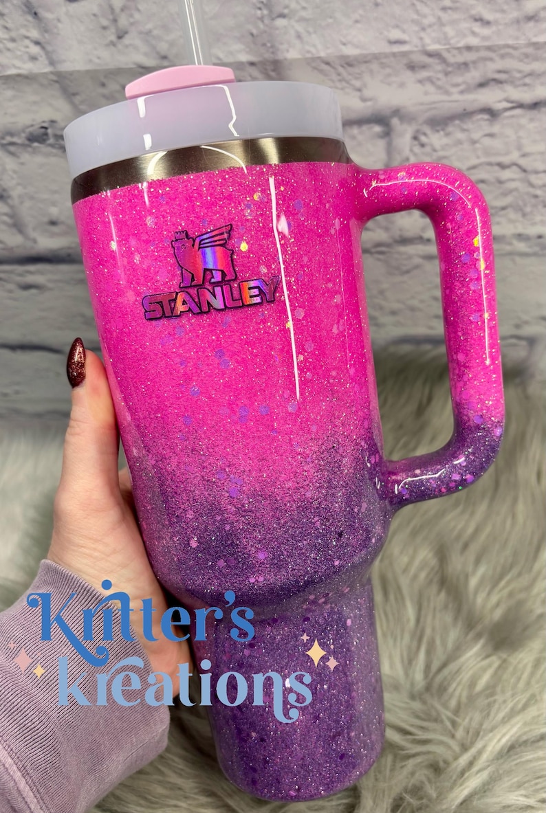 May include: A glittery Stanley tumbler in shades of pink and purple, with a handle and a straw. The tumbler features the Stanley logo and the text "Kitter's Kreations".