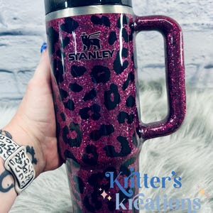 K&ouml;nnte beinhalten: Ein Stanley-Reisebecher mit Griff, mit einem pinkfarbenen Glitzer-Leopardenmuster. Der Becher hat einen schwarzen Deckel und Griff, mit dem Stanley-Logo. Der Text "Kritter's Kreations" ist ebenfalls auf dem Becher zu sehen.
