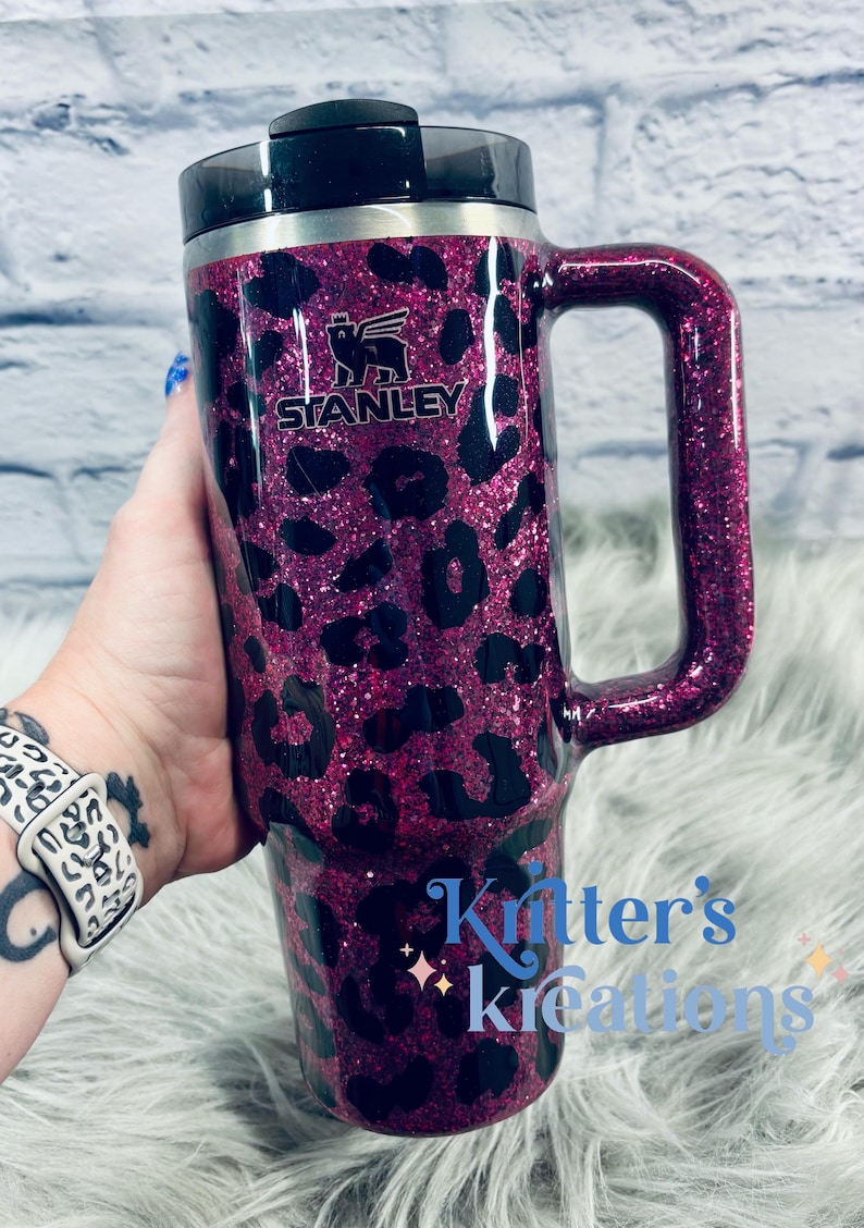 K&ouml;nnte beinhalten: Ein Stanley-Reisebecher mit Griff, mit einem pinkfarbenen Glitzer-Leopardenmuster. Der Becher hat einen schwarzen Deckel und Griff, mit dem Stanley-Logo. Der Text "Kritter's Kreations" ist ebenfalls auf dem Becher zu sehen.