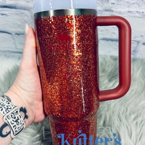 May include: A red glitter Stanley tumbler with a handle, white lid, and straw. The Stanley logo is visible on the front. The text "Knitter's Kreations" is at the bottom. The tumbler is held by a person.