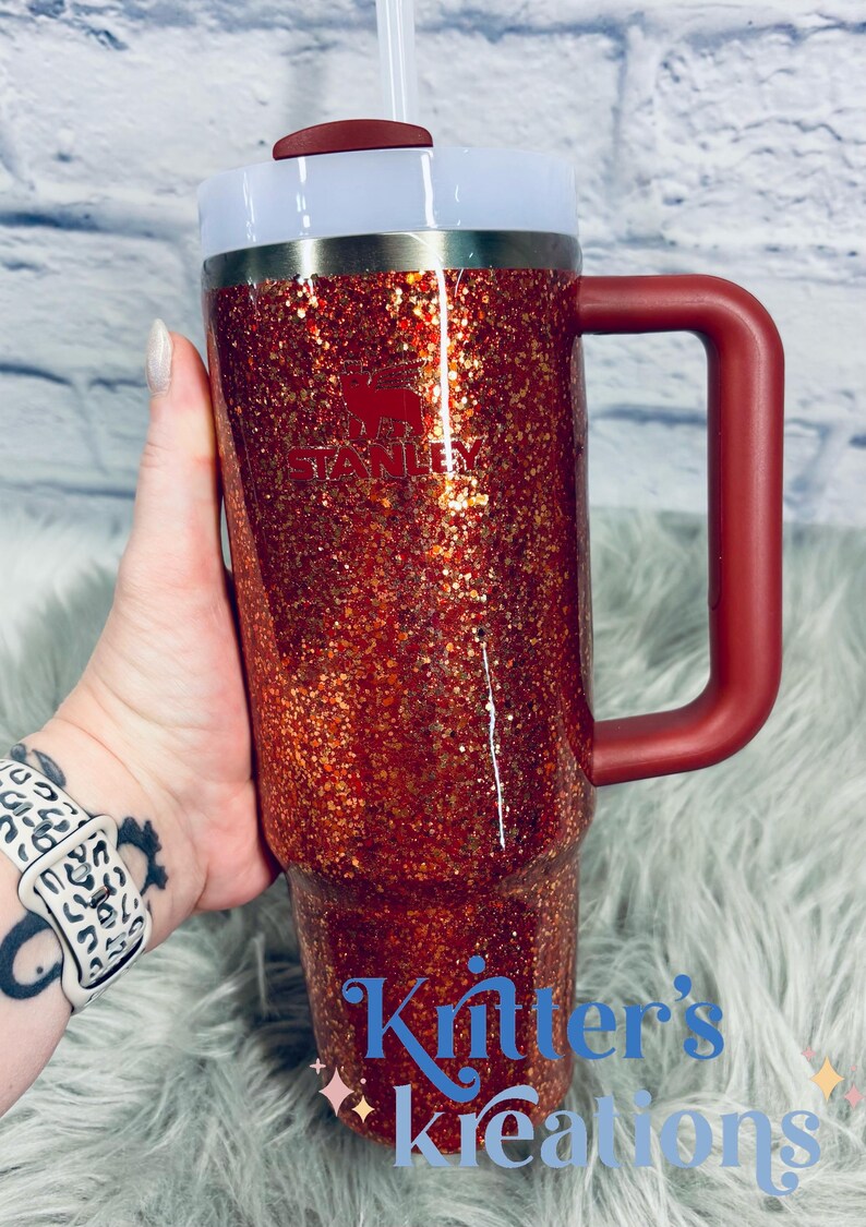 May include: A red glitter Stanley tumbler with a handle, white lid, and straw. The Stanley logo is visible on the front. The text "Knitter's Kreations" is at the bottom. The tumbler is held by a person.