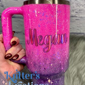 May include: A glittery pink and purple tumbler with a handle, featuring the name "Megan" in cursive. The tumbler has a light pink lid and a straw. The text "Kitter's Kreations" is also visible.
