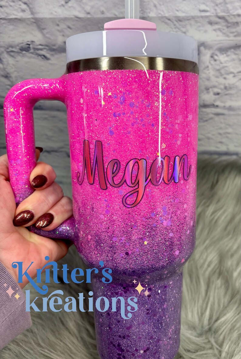 May include: A glittery pink and purple tumbler with a handle, featuring the name "Megan" in cursive. The tumbler has a light pink lid and a straw. The text "Kitter's Kreations" is also visible.