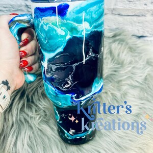 May include: A blue, black, and white marbled tumbler with a handle. The tumbler has a white lid and is decorated with a white swirl design. The tumbler is sitting on a fluffy, light blue surface. The text "Knitter's Kreations" is visible in the bottom right corner of the image.
