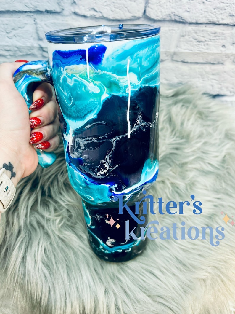May include: A blue, black, and white marbled tumbler with a handle. The tumbler has a white lid and is decorated with a white swirl design. The tumbler is sitting on a fluffy, light blue surface. The text "Knitter's Kreations" is visible in the bottom right corner of the image.