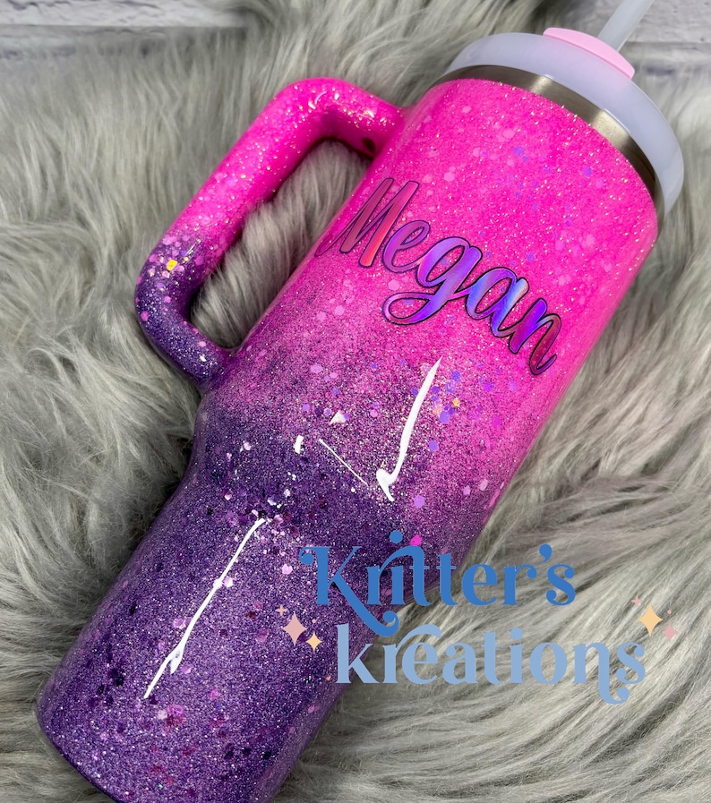 May include: A glitter-covered tumbler in shades of pink and purple, with a handle and a lid. The name "Megan" is written on the side. The words "Kritter's Kreations" are also visible. The tumbler is designed to keep drinks cold or hot.