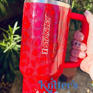 May include: A red Stanley tumbler with a black lid and handle. The tumbler has a glittery leopard print design and the Stanley logo. The text "Kritter's Kreations" is displayed in blue script on the bottom of the tumbler.